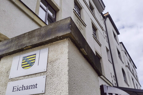 Schild mit Sachsen-Wappen und darunter der Aufschrift „Eichamt“. Im Hintergrund sieht man das helle Gebäude des Eichamtes Chemnitz.