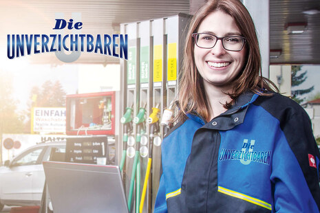 Eine Frau in Arbeitskleidung steht vor einer Tankstelle und lächelt ins Bild. Oben links im Bild und auf der Jacke der Frau steht das Logo von „Die Unverzichtbaren“.