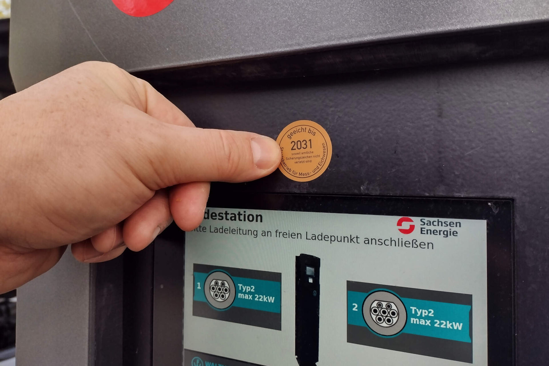 Zu sehen ist eine Hand, die ein Zusatzkennzeichen zur Eichung mit der Aufschrift „geeicht bis 2030, soweit amtliche Sicherungszeichen nicht verletzt sind“ rechts neben das Display einer E-Ladesäule klebt.
