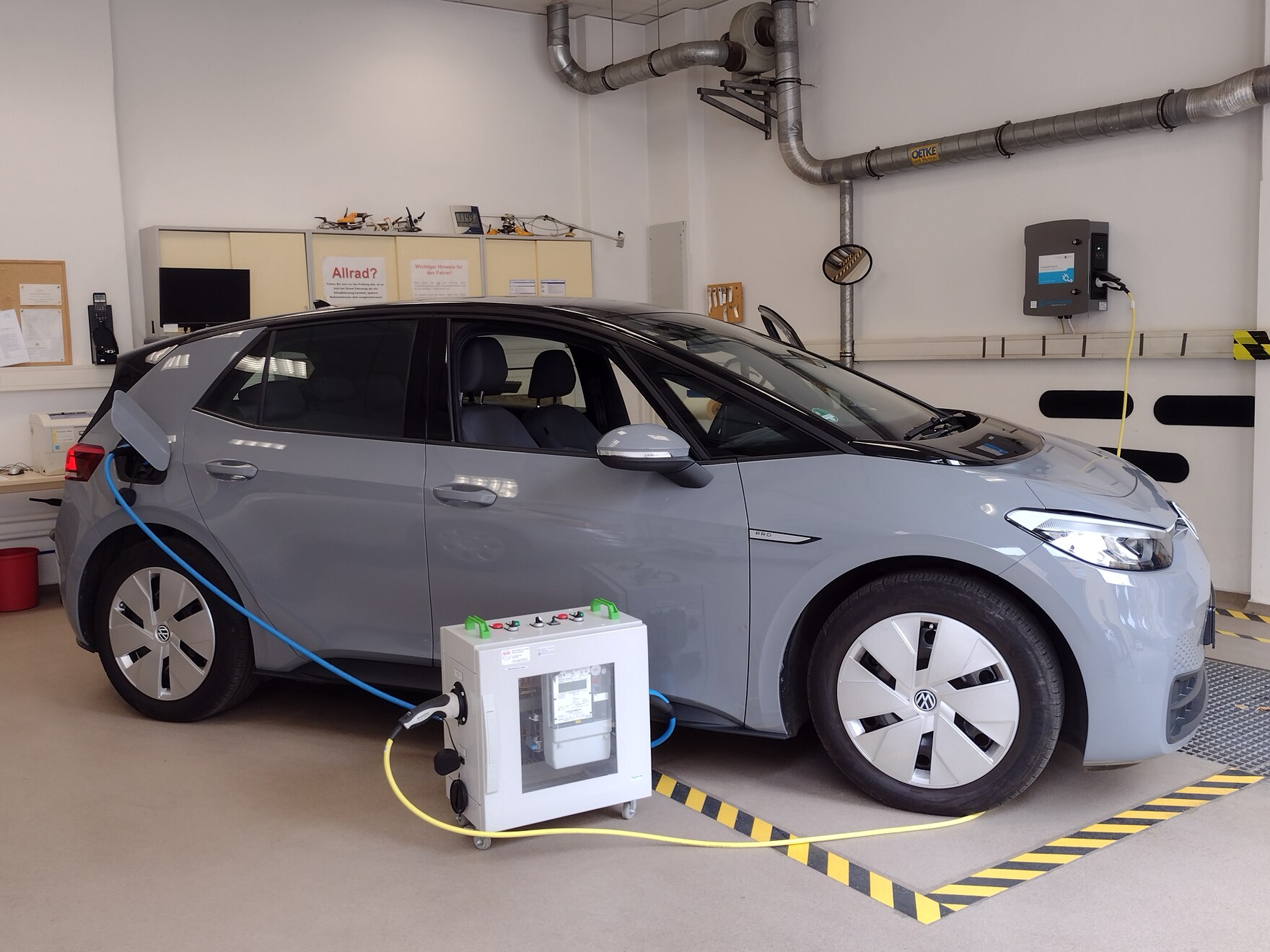 ID.3 Elektrofahrzeug, dass an einer Wallbox aufgeladen wird. Vor dem E-Fahrzeug steht ein Prüfgerät, welches zwischen Wallbox und Fahrzeug geschaltet ist und die tatsächliche Menge der Energie zum Aufladen des Fahrzeuges misst.