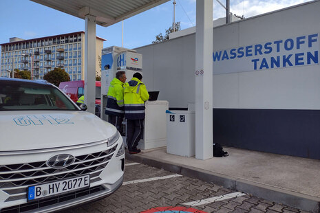 Zwei Personen in Arbeitskleidung an einer Tankstelle, eine tippt auf einem Laptop. An der Zapfsäule steht „H2“. Links ein Prüffahrzeug der Firma H2 Mobility zur Eichung der Wasserstofftankstelle. Im Hintergrund an der Wand: „Wasserstoff tanken“.