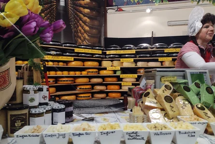 Verkaufsstand mit Käselaiben im Regal. Vorn stehen Schalen mit Käseproben. Eine Frau in holländischer Tracht überreicht einem Kunden die Ware. Daneben steht eine Waage.