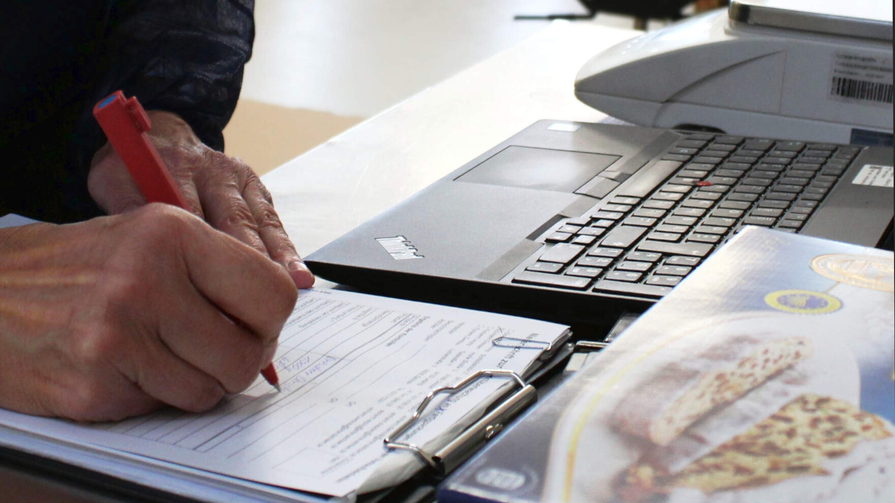 Im Vordergrund sieht man die Hände einer Person, die Daten in ein Prüfprotokoll schreiben. Daneben steht eine Fertigpackung mit Weihnachtsstollen. Im Hintergrund sieht man einen Laptop und eine Kontrollwaage.