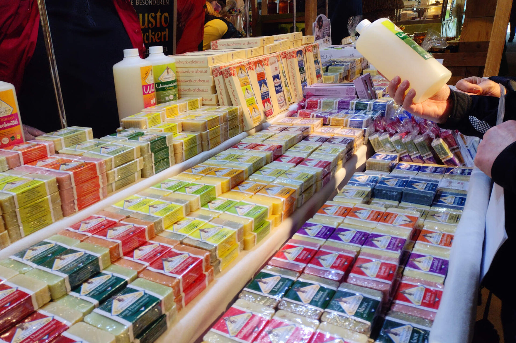 Marktüberwachung auf dem europäischen Bauernmarkt: Ein Verkaufsstand mit verschiedenen Seifenstücken in der Auslage. Am rechten Bildrand halten Hände eine Flasche hoch, während eine Person das Etikett liest.