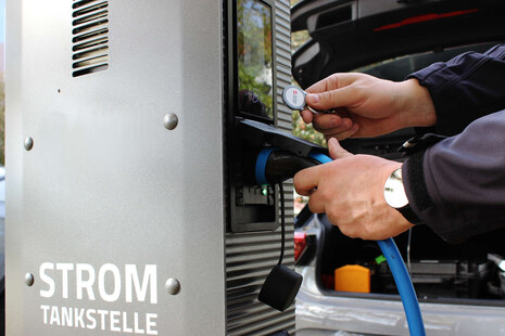 Ein Mann steht an einer E-Ladesäule und steckt gerade das Ladekabel seines Fahrzeuges an. Mit der rechten Hand hält er einen Chip an das Display der Ladesäule. Im Hintergrund sieht man das Elektrofahrzeug.