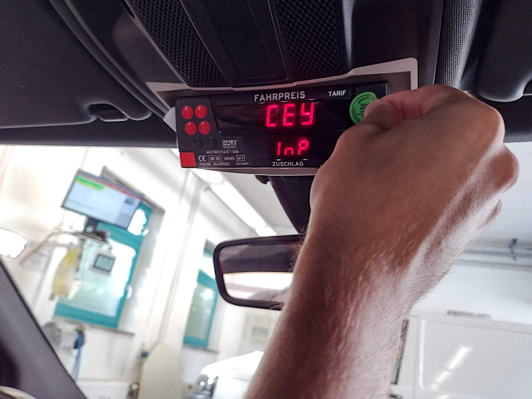 Im Innenraum eines Taxis klebt eine Hand ein grünes Kennzeichen zur Eichung auf den Taxameter. Das Taxameter hängt mittig über dem Rückspiegel. Rote zeichen leuchten im Bereich der Fahrpreisanzeige.