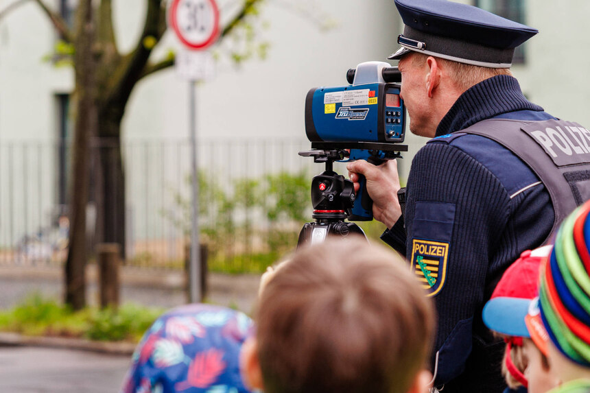 Ein Polizist steht an einer Straße und schaut durch ein mobiles Geschwindigkeitsmessgerät. Um ihn herum stehen Grundschulkinder. Im Hintergrund sieht man ein Verkehrsschild, das auf ein Tempolimit von 30 km/h hinweist.