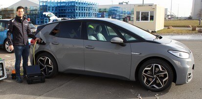 Ein Mann steht an einem grauen Elektrofahrzeug. Er steckt gerade den Ladestecker ein. Das Fahrzeug steht auf einem großen Parkplatz, im Hintergrund sieht man ein blau eingezäuntes Gebäude.