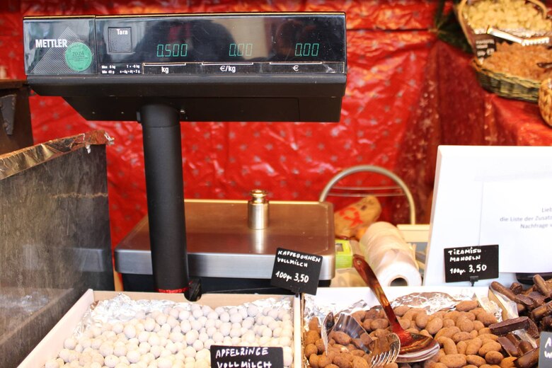 Auf einer Waage an einem Stand für schokolierte und karamellisierte Mandeln, Früchte und Kaffeebohnen steht ein 500-Gramm-Prüfgewicht. Die Anzeige zeigt 0,500 kg. Neben der Anzeige klebt ein grünes „geeicht bis 2024“-Kennzeichen.