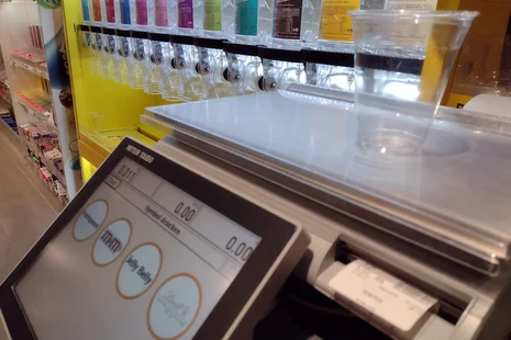 Foto einer Kunden-Wage im Supermarkt. Auf der Waage steht ein leerer Becher. Das Display zeigt 0 Gramm. Darunter sind vier Klickflächen zum Antippen, welche Ware man wiegen möchte. Im Hintergrund sieht verschiedene Süßigkeiten zum losen Verkauf.