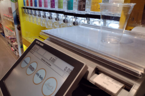 Foto einer Kunden-Wage im Supermarkt. Auf der Waage steht ein leerer Becher. Das Display zeigt 0 Gramm. Darunter sind vier Klickflächen zum Antippen, welche Ware man wiegen möchte. Im Hintergrund sieht verschiedene Süßigkeiten zum losen Verkauf.