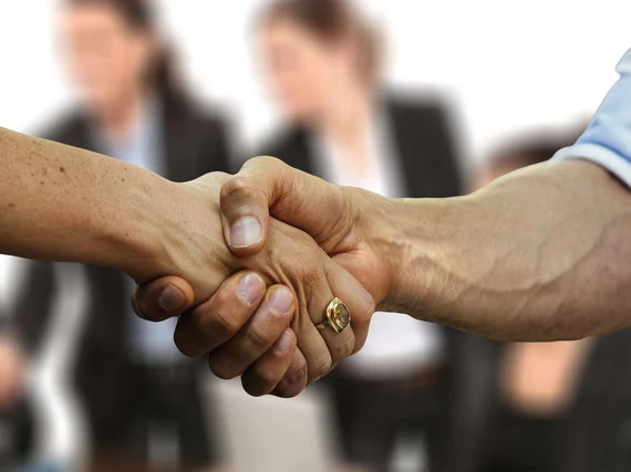 Im Vordergrund werden zwei Hände geschüttelt. Im Hintergrund sieht man unscharf mehrere Personen, wovon zwei Frauen in Businesskleidung aufgestanden sind und sich nach vorn beugen. Sie scheinen sich ebenfalls mit jemanden die Hände zu schütteln.