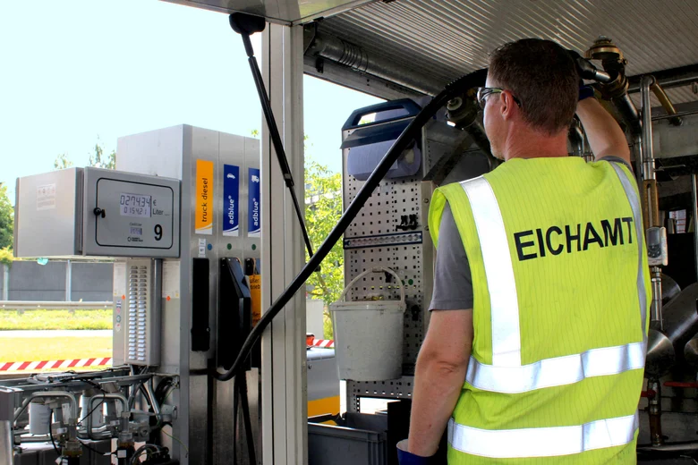 Ein Mann steht mit dem Rücken zur Kamera. Auf seiner Warnweste steht „Eichamt“. Er hält eine Zapfpistole in einen Kolben, der in seinem Prüffahrzeug verbaut ist und schaut zur Anzeige an der Zapfsäule. Die Anzeige lautet: 274,34 €, 154,21 Liter.