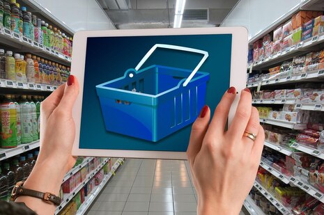 Im Vordergrund heben zwei Hände ein Tablet hoch, auf dessen Bildschirm ein Warenkorb abgebildet ist. Dahinter sieht man zwei Supermarktregale mit Flaschen und Verpackungen. Die Regale stehen sich gegenüber.